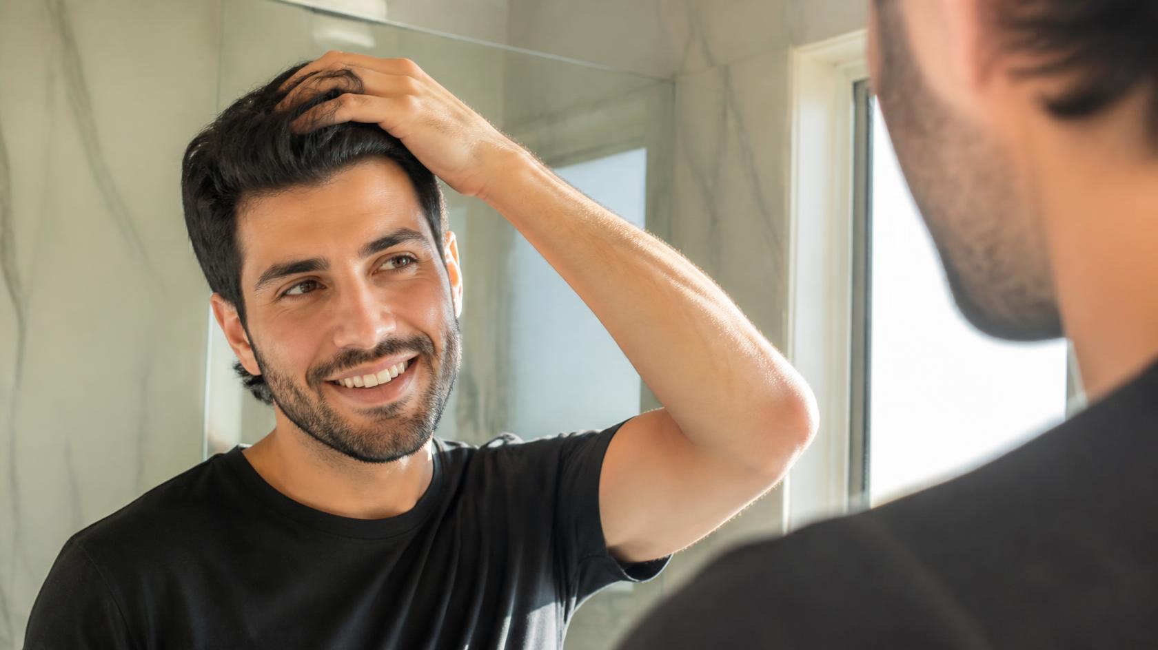 Smiling man confidently touching his healthy hair in sunlit bathroom mirror reflection.